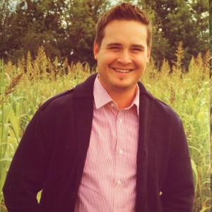 smiling man standing in front of a corn field in autumn wearing a red striped button up shirt and a navy blue cardigan